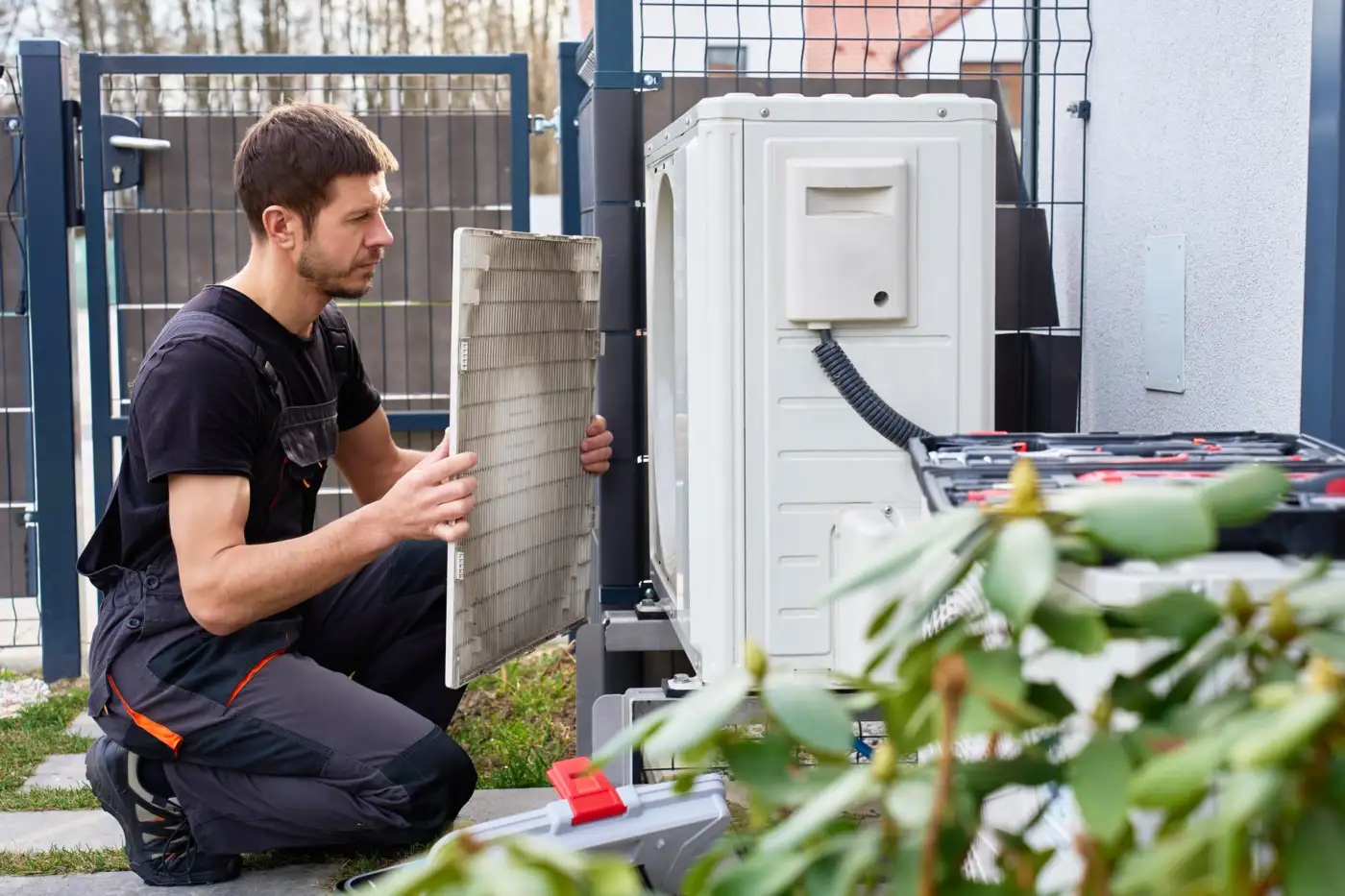 A heating engineer kneeling beside an outdoor heat pump unit at the side of a house