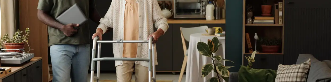 Older woman walking with a frame, supported by a carer in a home setting